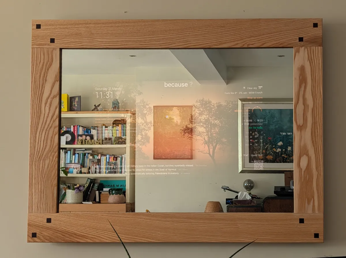 The Family Sentinel smart mirror mounted on a living room wall. A handmade ash frame with walnut corner splines surrounds two-way glass, through which a sunset landscape photo and floating weather and calendar information are visible. The room's bookshelves and furniture are reflected in the glass.